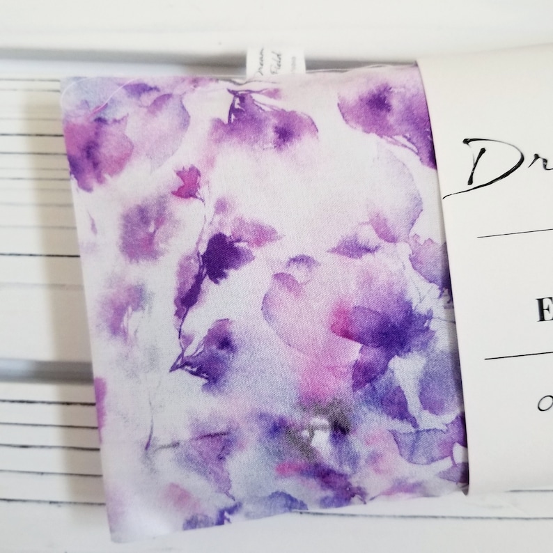 Op de afbeelding: Een gevouwen stoffen item met een aquarel bloemenpatroon in tinten paars en roze op een witte achtergrond. De stof is gedeeltelijk verpakt met een wit label met de tekst "Dream Field".