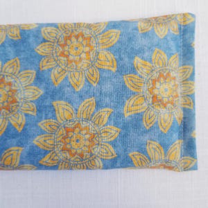 May include: A rectangular, blue fabric item with a repeating pattern of yellow and orange sunflowers. The sunflowers have detailed petals and centers. The item is likely a small pillow or sachet.
