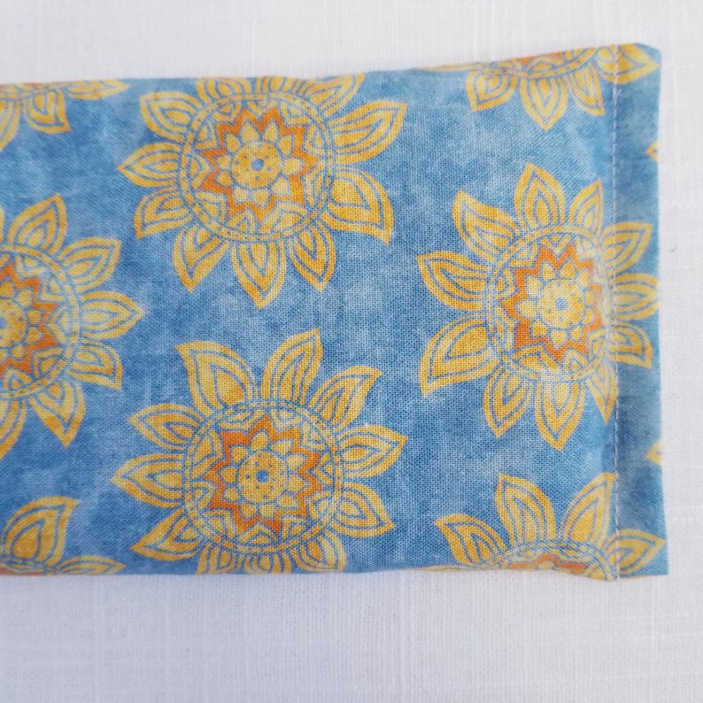 May include: A rectangular, blue fabric item with a repeating pattern of yellow and orange sunflowers. The sunflowers have detailed petals and centers. The item is likely a small pillow or sachet.