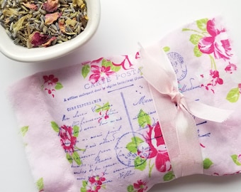 May include: A pink sachet with a floral print and a pink ribbon, next to a white bowl filled with dried herbs and flowers. The sachet has a vintage postcard design with text and floral accents.