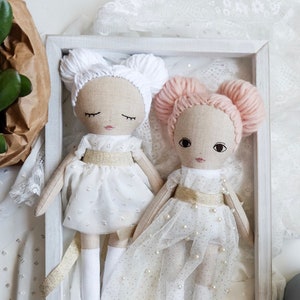 May include: Two handmade fabric dolls with white dresses and white yarn hair. The doll on the left has white yarn hair and the doll on the right has pink yarn hair. Both dolls are wearing white dresses with gold sashes and pearl embellishments.