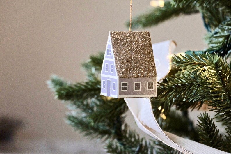 May include: A small, paper house ornament with a glitter roof. The house is light blue with white trim and windows. The ornament is hanging on a green Christmas tree.