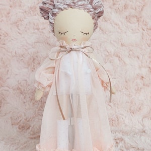May include: A pink and white doll with a crown on its head, wearing a pink tulle dress with a white bodice and a pink velvet bow. The doll has closed eyes and a pink blush on its cheeks.
