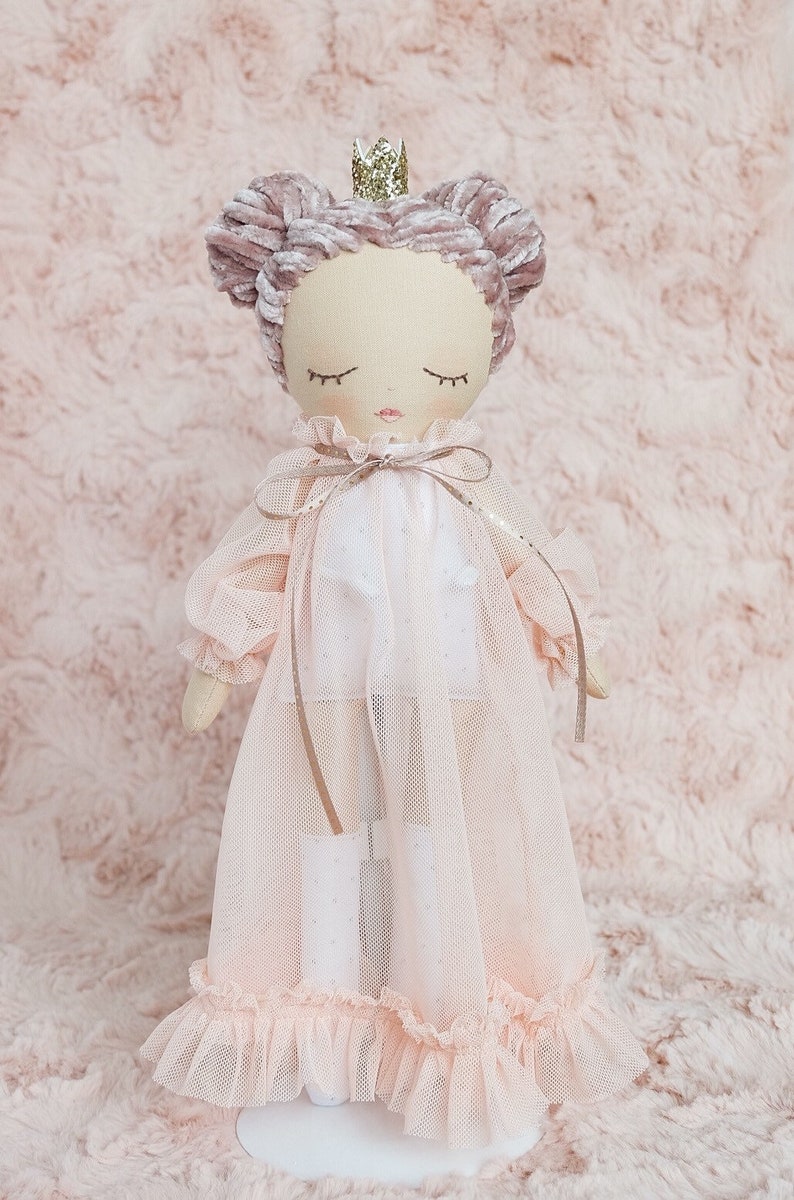 May include: A pink and white doll with a crown on its head, wearing a pink tulle dress with a white bodice and a pink velvet bow. The doll has closed eyes and a pink blush on its cheeks.
