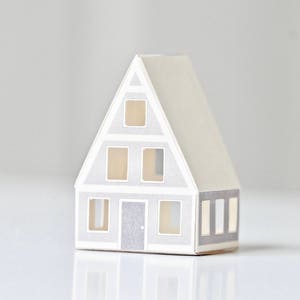 May include: A small paper model of a house with a gray roof and white walls. The house has five windows and a door. The windows and door are all gray. The house is sitting on a white surface.