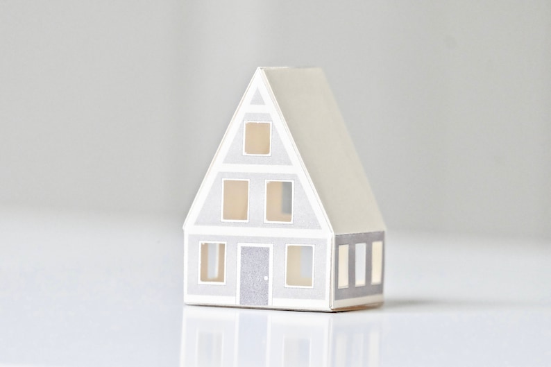 May include: A small paper model of a house with a gray roof and white walls. The house has five windows and a door. The windows and door are all gray. The house is sitting on a white surface.