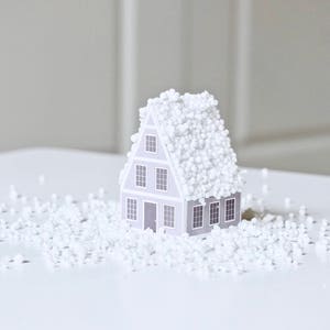 May include: A small gray house model covered in white packing peanuts. The house has four windows and a door. The house is sitting on a white surface.
