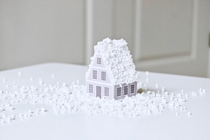 May include: A small gray house model covered in white packing peanuts. The house has four windows and a door. The house is sitting on a white surface.