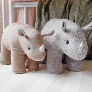 Può includere: Due giocattoli di peluche a forma di rinoceronte, uno grigio e uno marrone, sono seduti su una superficie bianca a righe. Il rinoceronte grigio ha una texture soffice, mentre il rinoceronte marrone ha una texture liscia, simile al lino.