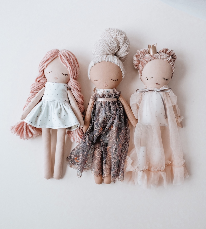 May include: Three handmade fabric dolls with different hairstyles and dresses. The doll on the left has pink hair and a white dress with polka dots. The doll in the middle has a brown dress with lace and a white bun. The doll on the right has a pink dress with a crown and a brown bun.
