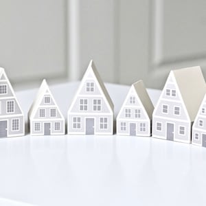 May include: Six gray paper houses of different sizes arranged in a row on a white surface. The houses have white trim and gray windows and doors. The houses are all facing the same direction.