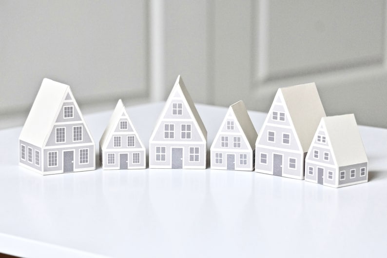 May include: Six gray paper houses of different sizes arranged in a row on a white surface. The houses have white trim and gray windows and doors. The houses are all facing the same direction.