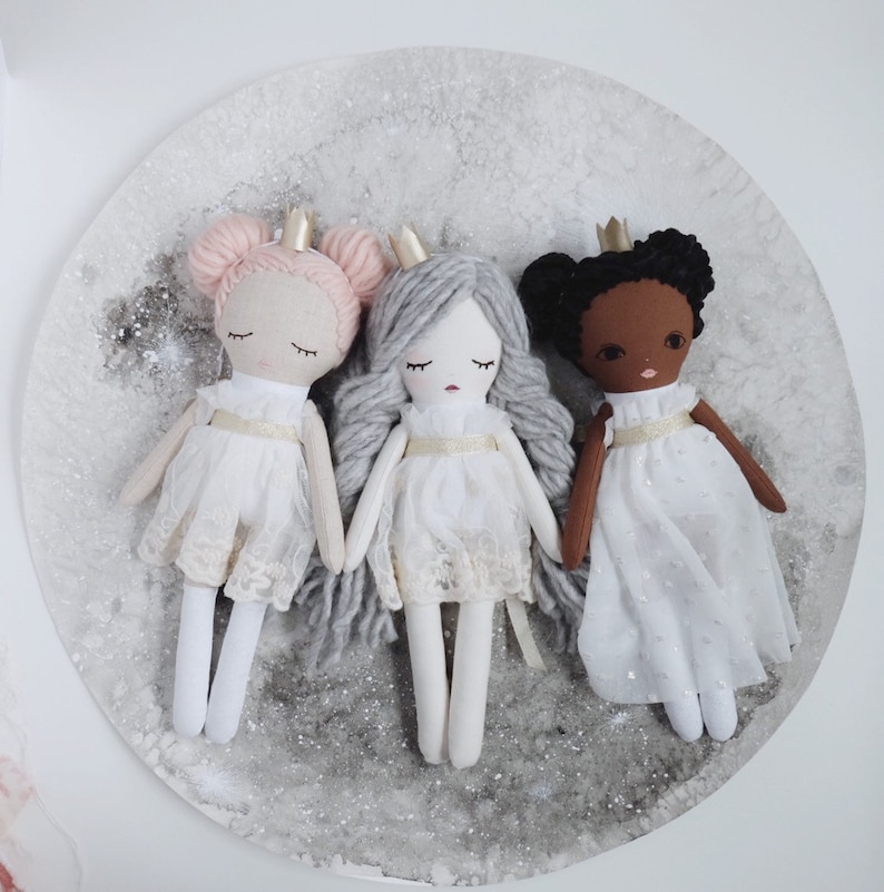 May include: Three handmade fabric dolls dressed as princesses. Each doll has a crown, a white dress with lace trim, and a different hair color: pink, gray, and brown.