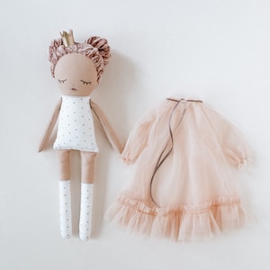 May include: A handmade doll with a white dress with black polka dots, pink hair, and a gold crown. The doll is wearing white socks and is holding a pink tulle dress.