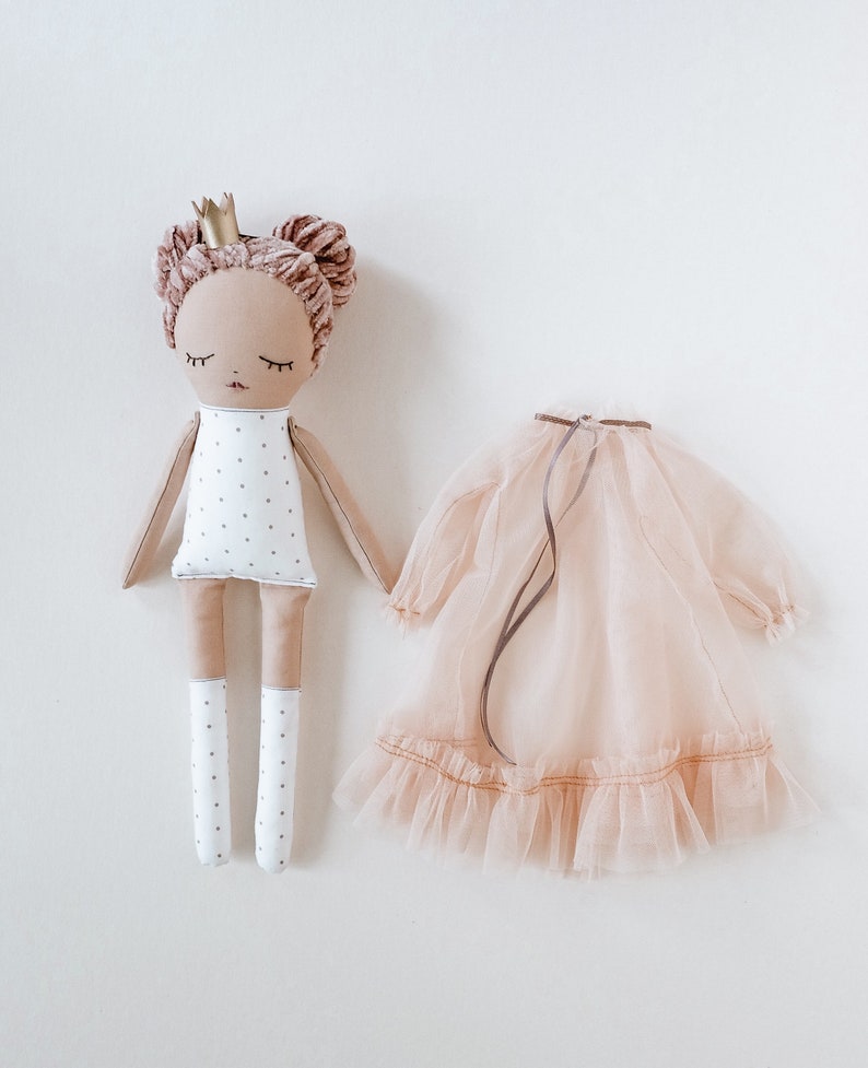 May include: A handmade doll with a white dress with black polka dots, pink hair, and a gold crown. The doll is wearing white socks and is holding a pink tulle dress.