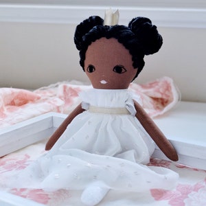 May include: A handmade doll with brown skin, black hair in two buns, and a white dress with a gold sash. The doll is wearing a gold crown and is sitting on a pink and white floral fabric.