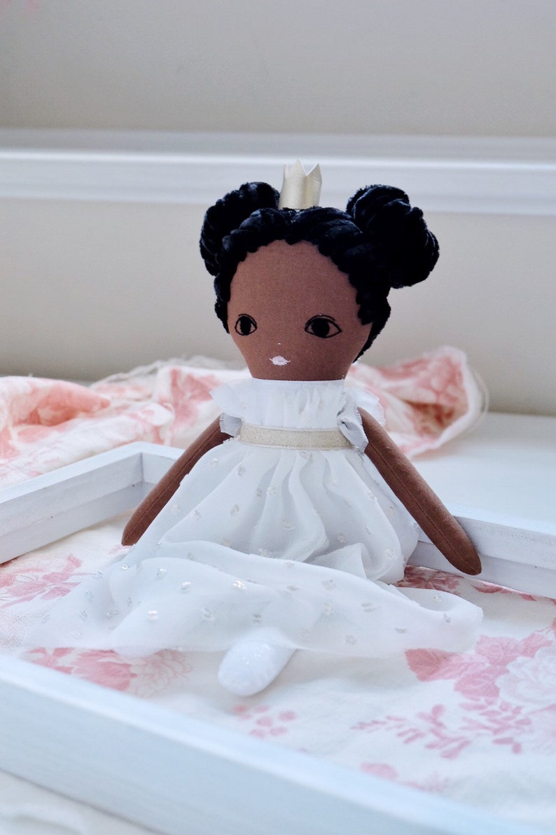 May include: A handmade doll with brown skin, black hair in two buns, and a white dress with a gold sash. The doll is wearing a gold crown and is sitting on a pink and white floral fabric.