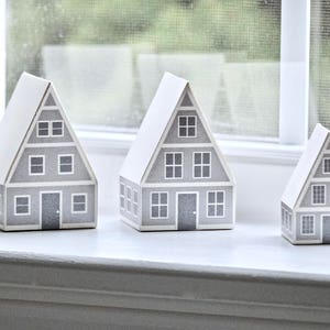 May include: Five gray paper houses with white trim and windows, arranged on a white windowsill.