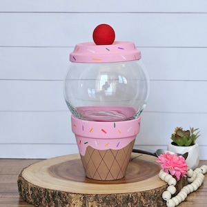 May include: A pink and brown ice cream cone shaped glass jar with a red cherry on top. The jar is decorated with sprinkles and a waffle cone pattern.