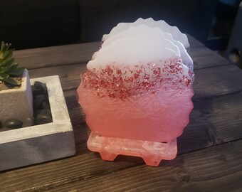 May include: Set of four pink and white resin coasters with a glitter finish. The coasters are stacked on a small stand.