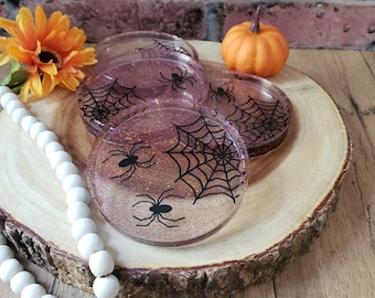 May include: Four pink glitter coasters with black spiderwebs and spiders. The coasters are on a wooden surface with a small orange pumpkin.