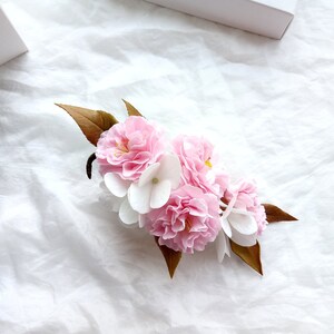 May include: A cluster of pink and white paper flowers with brown leaves. The flowers are arranged in a circular shape and are set against a white background.