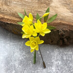 May include: A yellow flower boutonniere with a gold pin. The flower is made of fabric and has a realistic appearance.