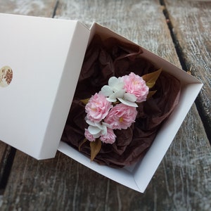 May include: A white gift box with a brown paper filler and a bouquet of pink and white paper flowers inside.