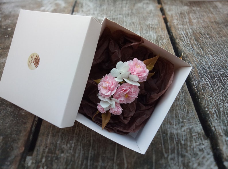 May include: A white gift box with a brown paper filler and a bouquet of pink and white paper flowers inside.