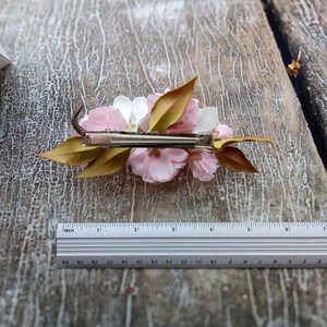 May include: A white and pink flower hair clip with green leaves. The clip is made of metal and has a spring mechanism.