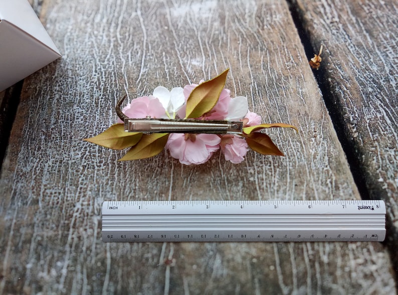 May include: A white and pink flower hair clip with green leaves. The clip is made of metal and has a spring mechanism.
