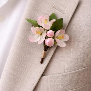May include: A floral boutonniere featuring delicate pink blossoms with yellow centers and green leaves. The arrangement is attached to a brown stem wrapped with twine. The boutonniere is pinned to a light gray suit jacket.