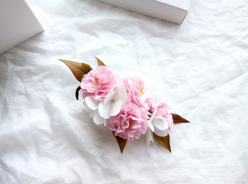 May include: A cluster of pink and white paper flowers with brown leaves. The flowers are arranged in a circular shape and are set against a white background.