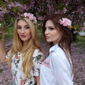 May include: Two women wearing floral crowns and floral hair clips stand in a field of pink flower petals. One woman is wearing a white floral print shirt and green pants. The other woman is wearing a white shirt with embroidered flowers and a pink skirt.