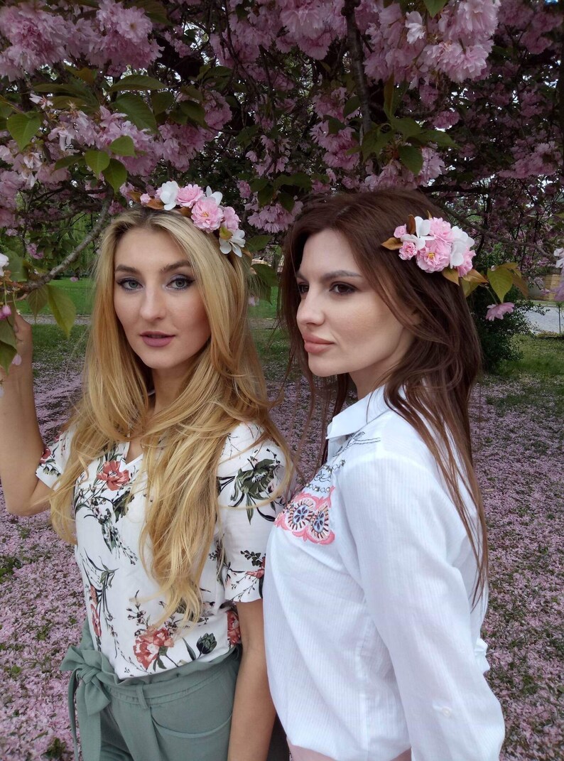 May include: Two women wearing floral crowns and floral hair clips stand in a field of pink flower petals. One woman is wearing a white floral print shirt and green pants. The other woman is wearing a white shirt with embroidered flowers and a pink skirt.