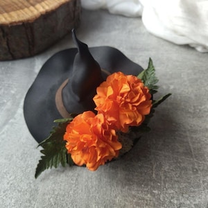 Pode incluir: Um chapéu de bruxa preto com uma borda marrom e duas flores de cravo-da-índia laranja.