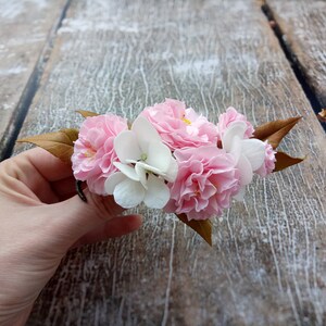 May include: A bouquet of pink and white fabric flowers with brown leaves. The flowers are arranged in a cluster and are held in a hand.