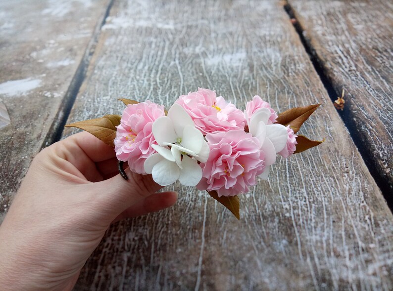 May include: A bouquet of pink and white fabric flowers with brown leaves. The flowers are arranged in a cluster and are held in a hand.