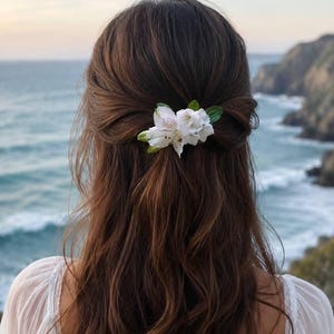 May include: A hair accessory featuring white alstroemeria flowers with subtle purple speckles and green leaves, secured in long, wavy brown hair. The floral piece is positioned at the back of the head, holding a section of hair in a half-up style.
