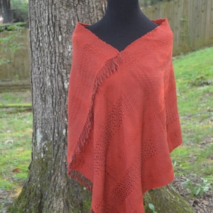 May include: A rust-coloured woven shawl with fringe on the edges. The shawl is draped over a black mannequin.