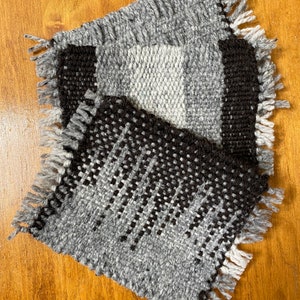 Puede incluir: Tres posavasos tejidos de color gris y negro con bordes con flecos. Los posavasos están apilados uno encima del otro.