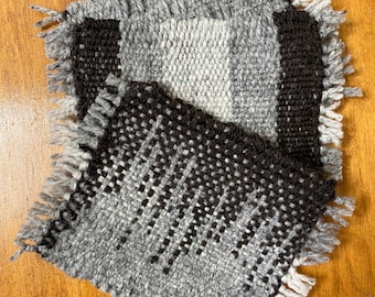 May include: Three woven gray and black coasters with fringed edges. The coasters are stacked on top of each other.