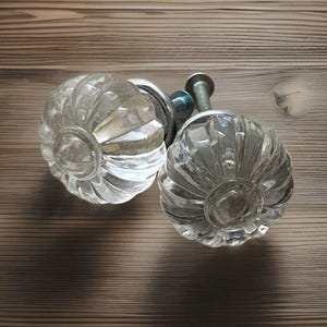Puede incluir: Dos pomos de armario de cristal transparente con un diseño redondeado y acanalado. Los pomos tienen una estética vintage y están montados sobre herrajes plateados. Los pomos están colocados sobre una superficie de madera marrón claro.