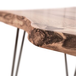May include: A close-up of a wooden table with a live edge top and black metal hairpin legs. The wood has a natural grain pattern and a dark brown colour.