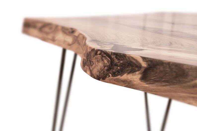 May include: A close-up of a wooden table with a live edge top and black metal hairpin legs. The wood has a natural grain pattern and a dark brown colour.