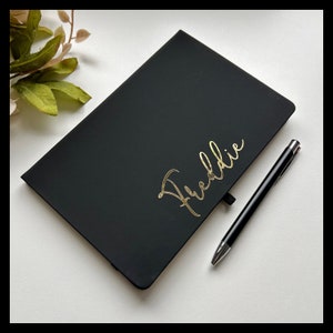 May include: A black notebook with the name "Freddie" written in gold lettering. A black pen is lying next to the notebook.