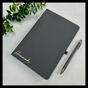 May include: A charcoal grey notebook with the name "Hannah" in gold script, alongside a matching pen. The notebook is closed and has a ribbon bookmark. Green foliage is in the background.