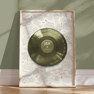 May include: A framed print with a gold-coloured vinyl record at its centre, inscribed with the initials "J&B" and the words "Happily ever after." The background is cream-coloured paper covered in handwritten text. The frame is light wood.