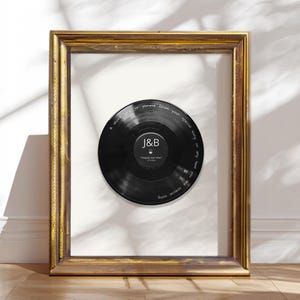 May include: A black vinyl record displayed inside a rectangular gold frame. The record features a central label with the letters J&B and the text "happily ever after" with a date. White text is etched along the outer edge of the vinyl record.