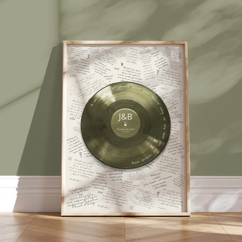 May include: A framed print with a gold-coloured vinyl record at its centre, inscribed with the initials "J&B" and the words "Happily ever after." The background is cream-coloured paper covered in handwritten text. The frame is light wood.